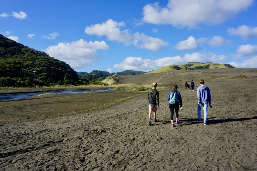 Three people in foreground walking towards black sand dunes, with stream running along left of picture - Bethells Beach, New Zealand