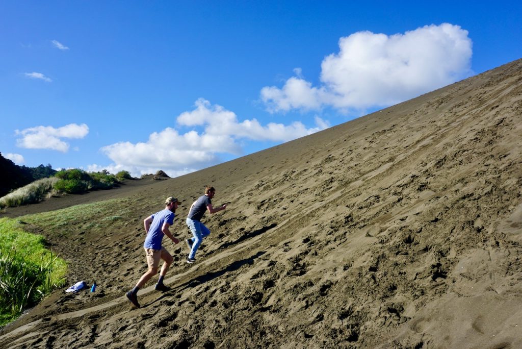 Two men running up steep black sand dune - Bethells Beach, New Zealand