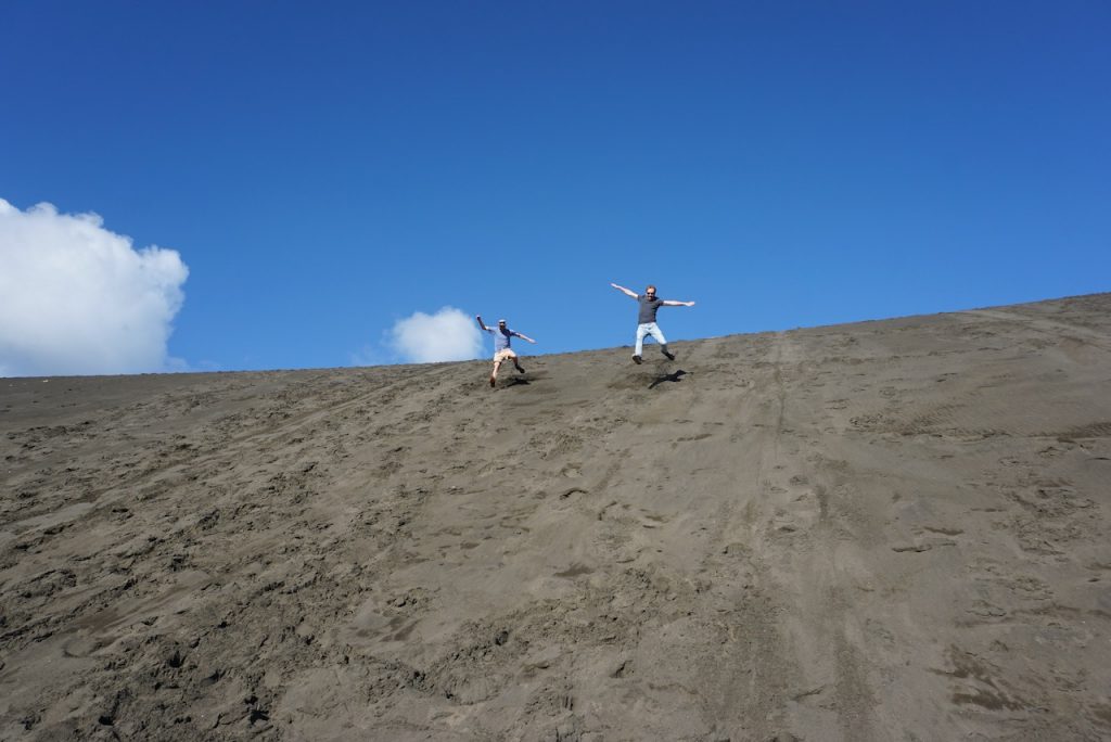 Two men hurtling down steep black sand dune with blue sky behind - Bethells Beach, New Zealand