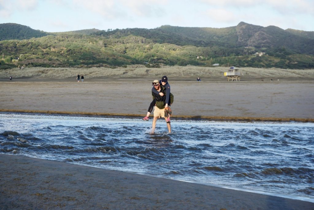 Man carries woman on his back across stream at black sand beach, Bethells Beach, New Zealand
