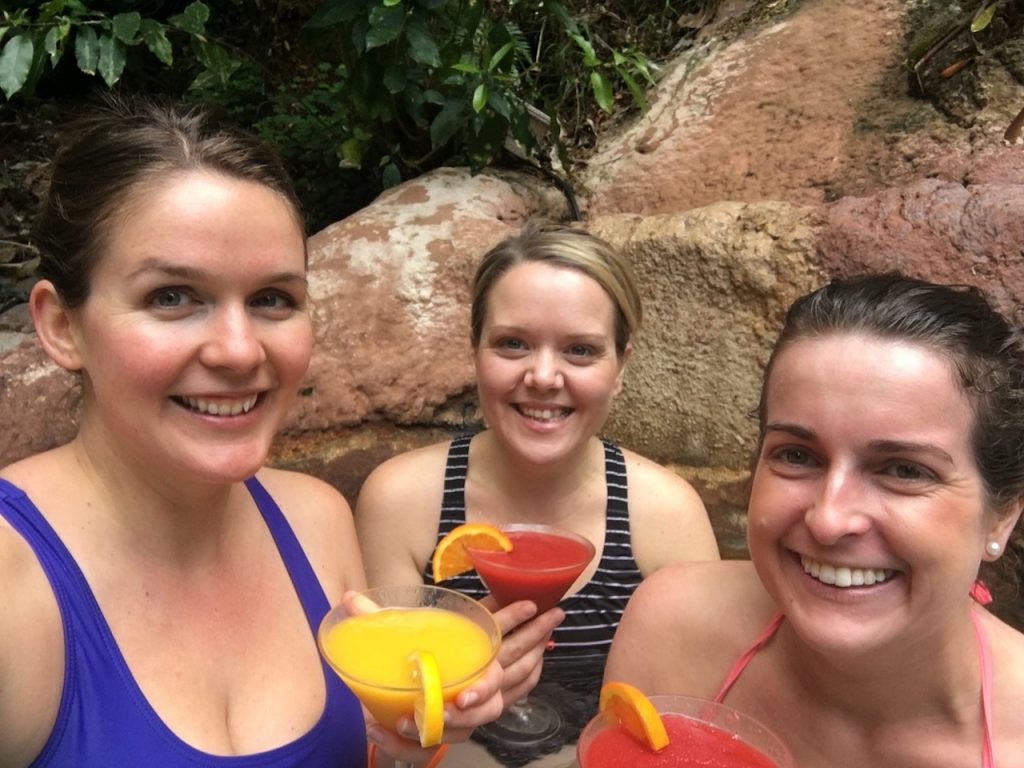 Three women in swim suits in hot pool holding brightly coloured cocktails